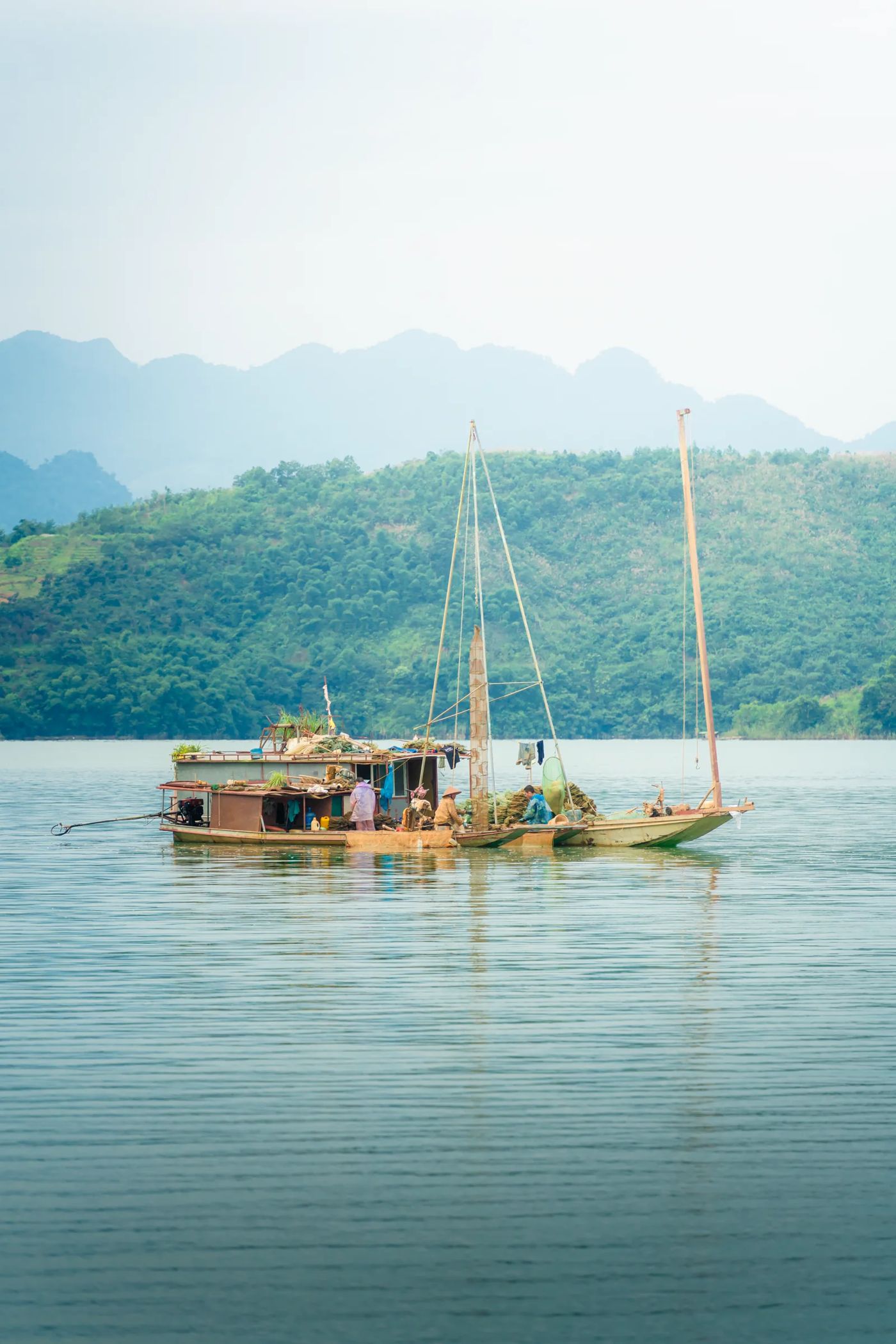 Thuyền buồm của người dân trên mặt hồ phẳng lặng, núi mờ phía sau